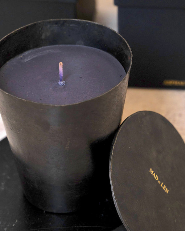 Black candle in a holder with a lid on a light wooden surface, with black boxes in the background.
